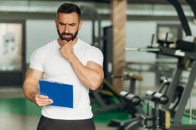 Muscular handsome trainer looking at fitness plan on clipboard for working out in the fitness gym