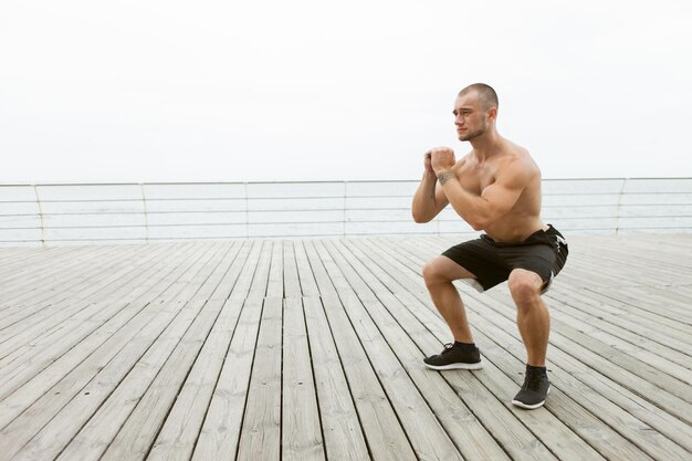 Muscular male athlete with naked torso doing squats exercise on the beach