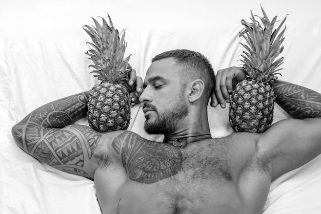 Muscular man hold pineapple fruit on biceps brutal sportsman relax in bed steroids sexy ab of tattoo