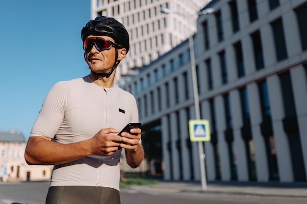 Muscular man in sport clothes helmet and glasses looking aside while standing on street with modern smartphone in hands Modern building on background