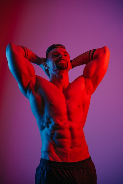 A muscular man with a beard is posing with arms behind his head under the blue and red light