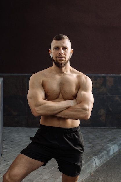 Muscular man with naked torso wearing an earphones is posing against the concrete wall outdoors