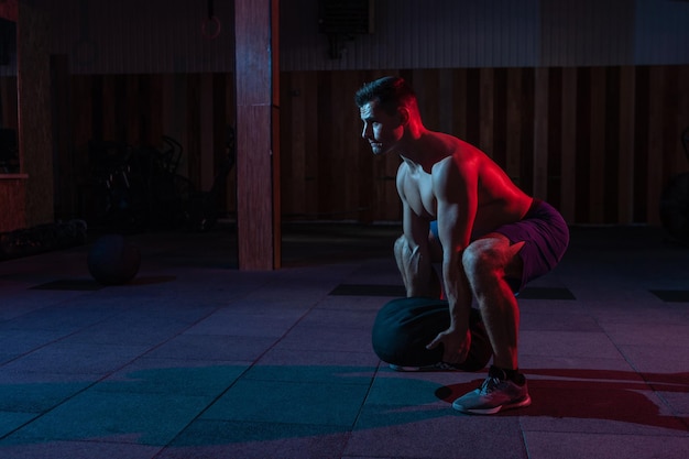 A muscular powerful man with a naked torso exercises with a heavy bag in a red blue neon light Cross workout in a modern gym