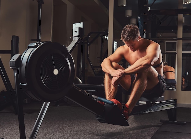 A muscular shirtless athlete resting after a hard workout while sits on the rowing machine in the gym.