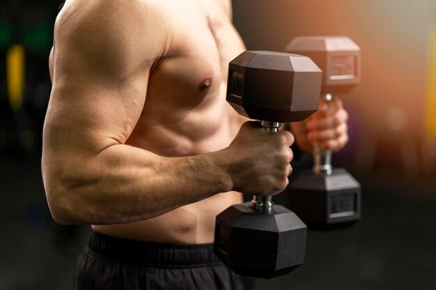 Muscular strong man with a bare torso holding dumbbells training in gym closeup