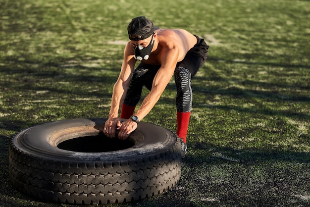 Photo muscular young male with mask doing exercises with big tire outdoors in stadium shirtless sportsman doing hard workout healthy lifestyle people and sport concept