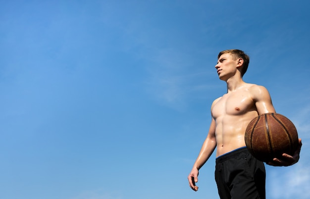 Muscular young sportsman with basketball ball outdoors against sky background.