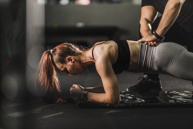 Muscular young woman working out with personal trainer at the gym. She is doing plank exercises.