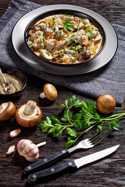 Mushroom and calf liver stroganoff in a black bowl on a dark rustic wooden table with ingredients and spices