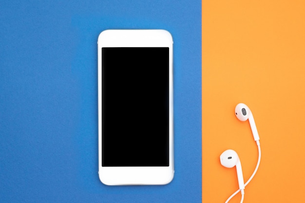 Music, gadgets, music lover. White smartphone and headphones on the orange and blue background with headphones. View from above. Flat lay, top view.