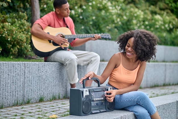 Music lovers. Dark-skinned guy playing guitar and smiling girl with tape recorder sitting in park on fine day