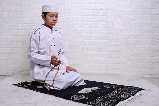 Muslim kid wearing white rob and skullcap is sitting and holding prayer beads at prayer mat