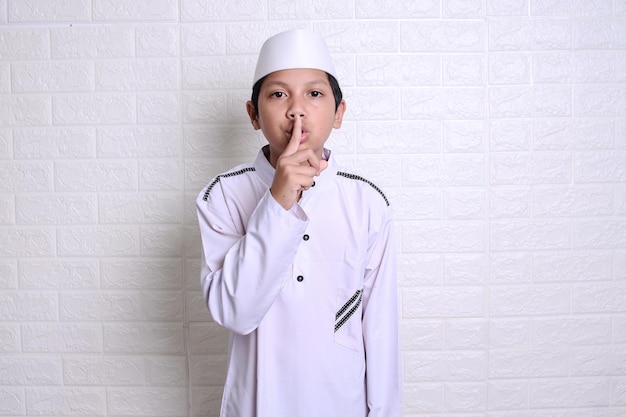 muslim kid in white rob and skullcap gesturing maintain silence on white background. Be quite please