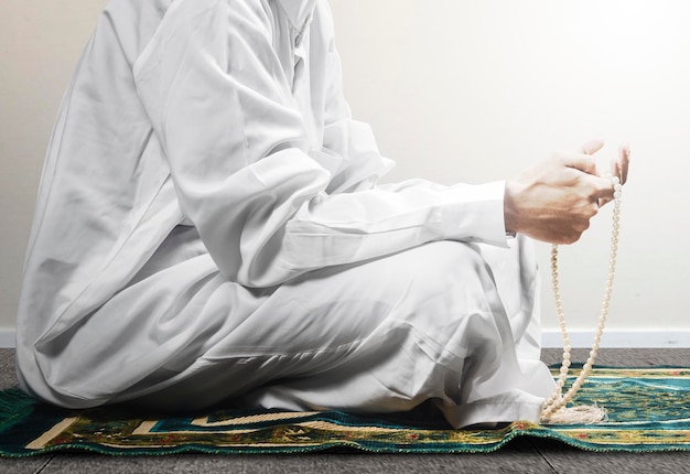 Muslim man praying with prayer beads on his hands while sitting on the prayer rug with white wall background