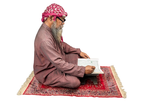 Muslim man with a beard wearing keffiyeh with agal sitting and reading the Quran on the prayer rug