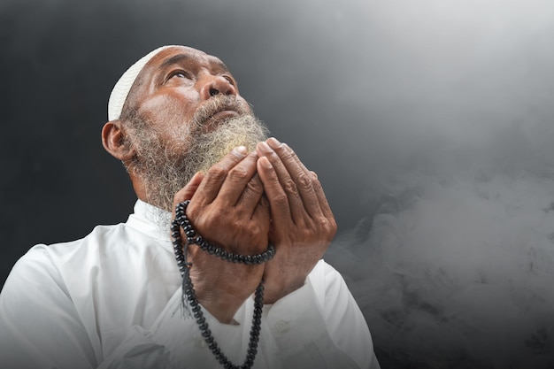 Muslim man with a beard wearing a white cap praying with prayer beads on his hands