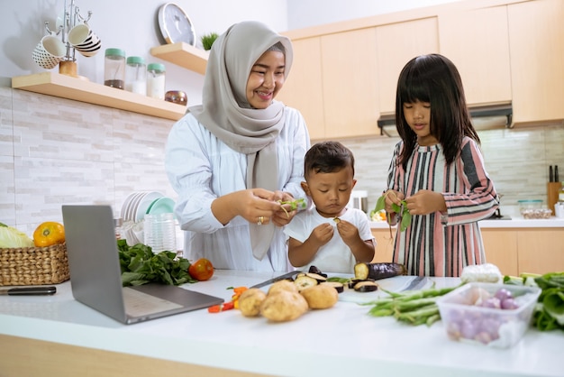 Muslim mother watching cooking video on laptop and making dinner with her two children in the kitchen together