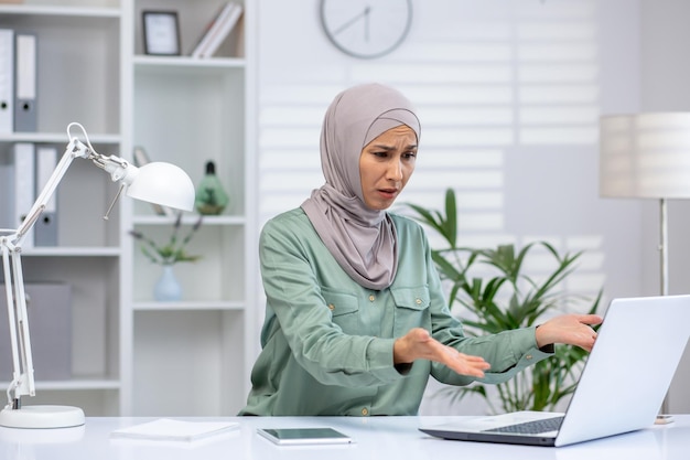 A muslim woman in a hijab appears stressed and confused while sitting at her desk dealing with an