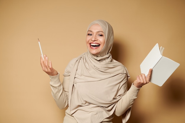 Muslim woman in hijab holding a notepad and pencil and laughing while posing to front on beige surface with copy space