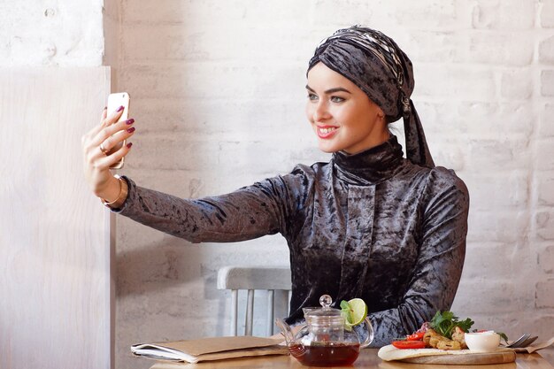 Photo muslim woman sitting in cafe takes pictures of herself on the phone.