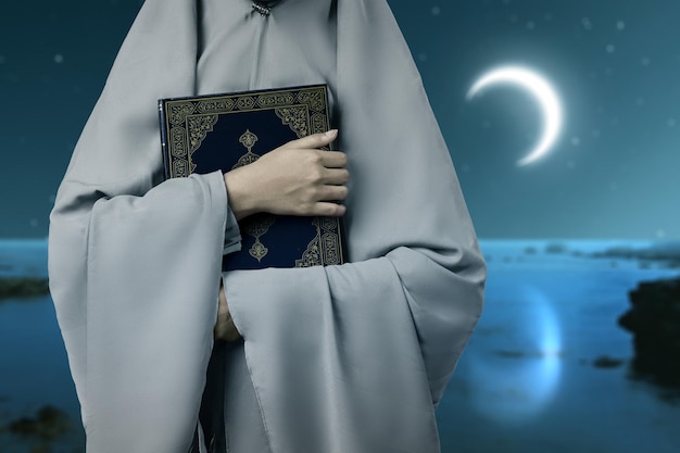 Muslim woman in a veil standing and holding the Quran with the night scene background