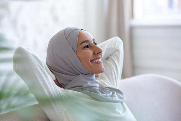 Muslim young female student in hijab sitting on sofa at home hands behind head and resting smiling