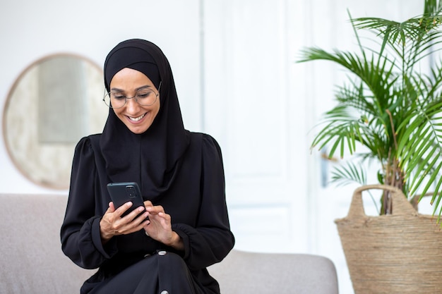 Muslim young woman in black hijab and burqa sitting on sofa at home and using mobile phone smiling