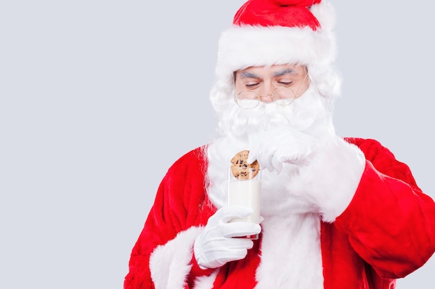 My favorite cookies! Traditional Santa Claus holding cookie and glass with milk while standing against grey background