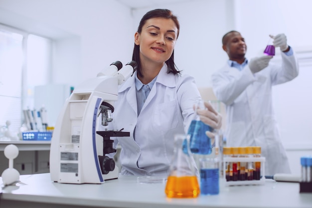 My lab. Cheerful skilled scientist working with the tubes and her colleague standing in the background