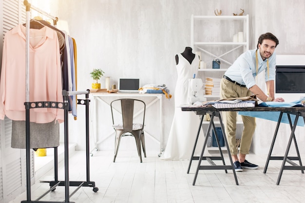 My own office. Cheerful skilled dressmaker standing at the table and making a new dress