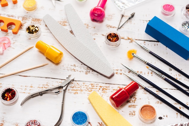 Nail files, scissors, pliers, sequins and nail polishes on a white wooden background.