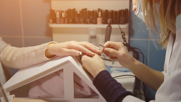 Nail master treats the cuticle of a young client with manicure forceps in a beauty salon, sunlight.