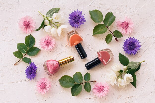 Nail polish bottles with jasmine flowers, cornflowers and green leaves on white surface