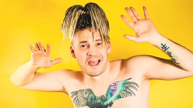 Naked handsome energetic man with tattoo on chest dancing looking at camera smiling on yellow background Enthusiastic male with dreadlocks dancing with good mood gesturing hands in studio