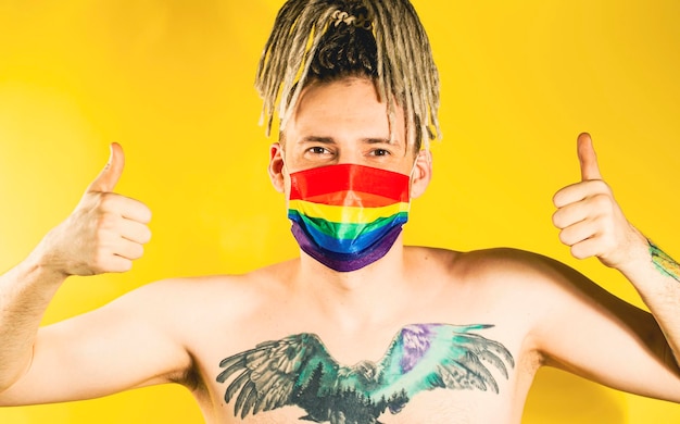 Naked handsome young man in LGBT mask showing gesture of like Attractive gay guy with blonde dreadlocks in protective rainbow mask tattoo on chest showing thumbs up on yellow background