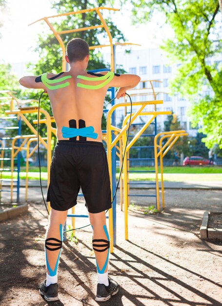 Naked muscular man training with fitness rope at sports ground. Back view of young unrecognizable bodybuilder with elastic kinesiology taping on body training outdoors. Rehabilitation concept.