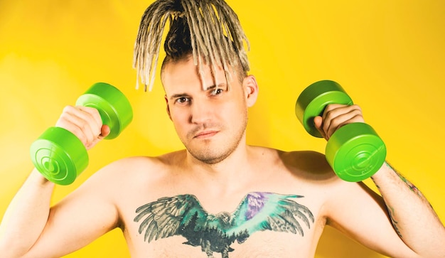 Naked strong man lifting dumbbells training muscles Determined active guy with blonde dreadlocks tattoo on chest doing sport weight training on yellow background in studio