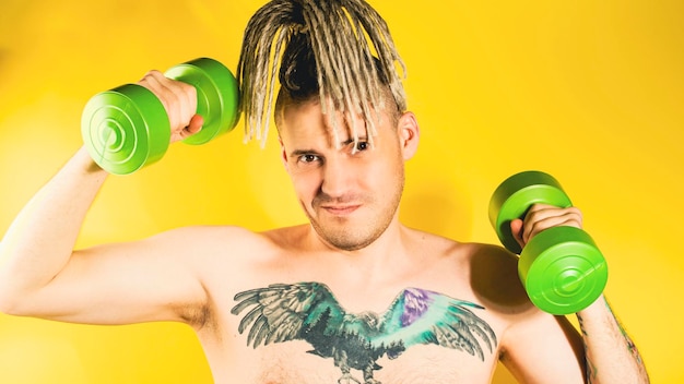 Naked strong man lifting dumbbells training muscles Determined active guy with blonde dreadlocks tattoo on chest doing sport weight training on yellow background in studio