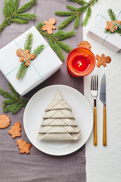 Napkin in the form of a Christmas tree on a plate on white tablecloth with gifts and decorations with fir sprigs and gingerbread cookies