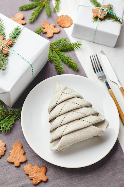 Napkin in the form of a Christmas tree on a plate on white tablecloth with gifts and decorations with fir sprigs and gingerbread cookies