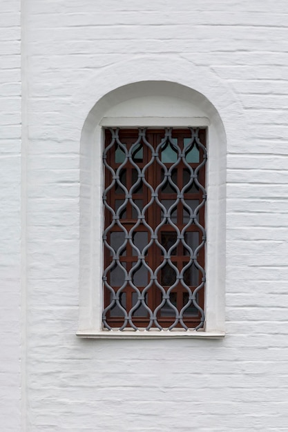 A narrow vertical window with bars in the white brick wall of the fortress Closeup