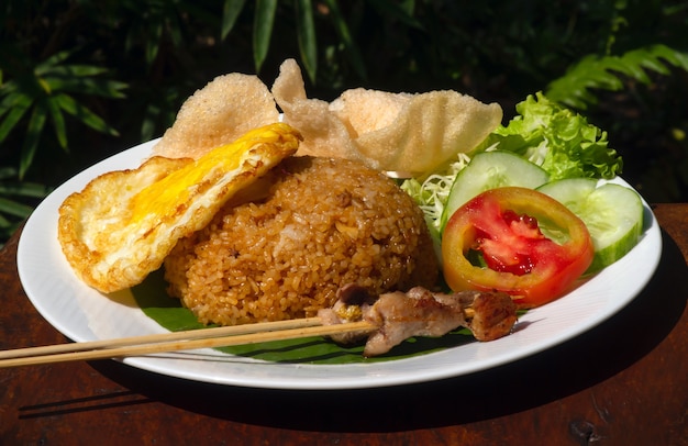 Nasi Goreng, fried rice, traditional Indonesian food, with chicken egg and vegetables on a white plate. Indonesian cuisine.