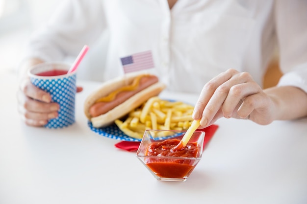 national holidays, celebration, food and patriotism concept - close up of woman eating french fries with hot dog and drinking juice from paper cup at 4th july at party on american independence day