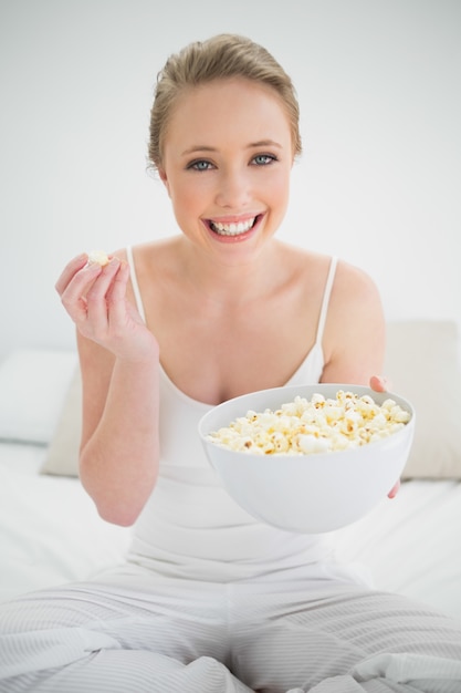 Natural cheerful blonde holding bowl of popcorn on bed