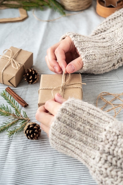 Natural Christmas background, craft gift boxes, cones, Christmas tree on the table, close-up, retro style. A girl in a sweater is packing gifts. The happy atmosphere of the holiday.