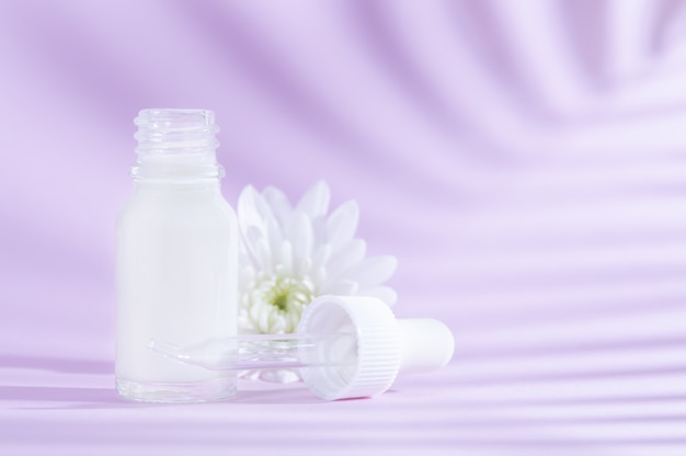 Natural cosmetics: serum with dropper and white flower on lilac background with shadow.