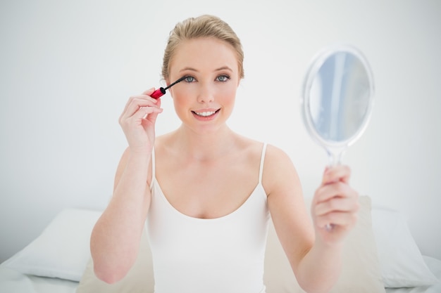 Natural smiling blonde holding mirror and applying mascara