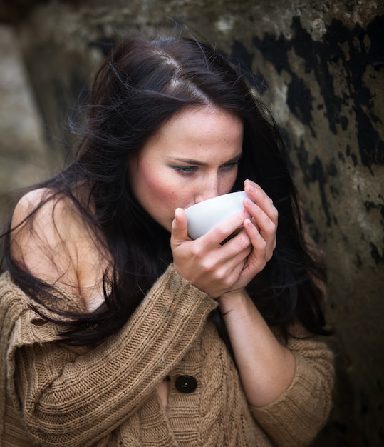 Nature, beauty, youth and healthy lifestyle concept. Young cute girl in a knitted sweater against a background of an old concrete wall drinking tea from a white cup