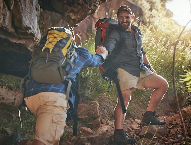 Nature fitness and hiking friends climbing up rocks on a mountain hike trail during spring Fit men holding hands to help and support trekking up stones with a backpack on adventure health workout