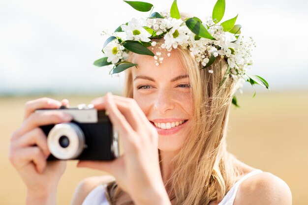 nature, summer holidays, vacation and people concept - happy woman in wreath of flowers taking picture with film camera outdoors